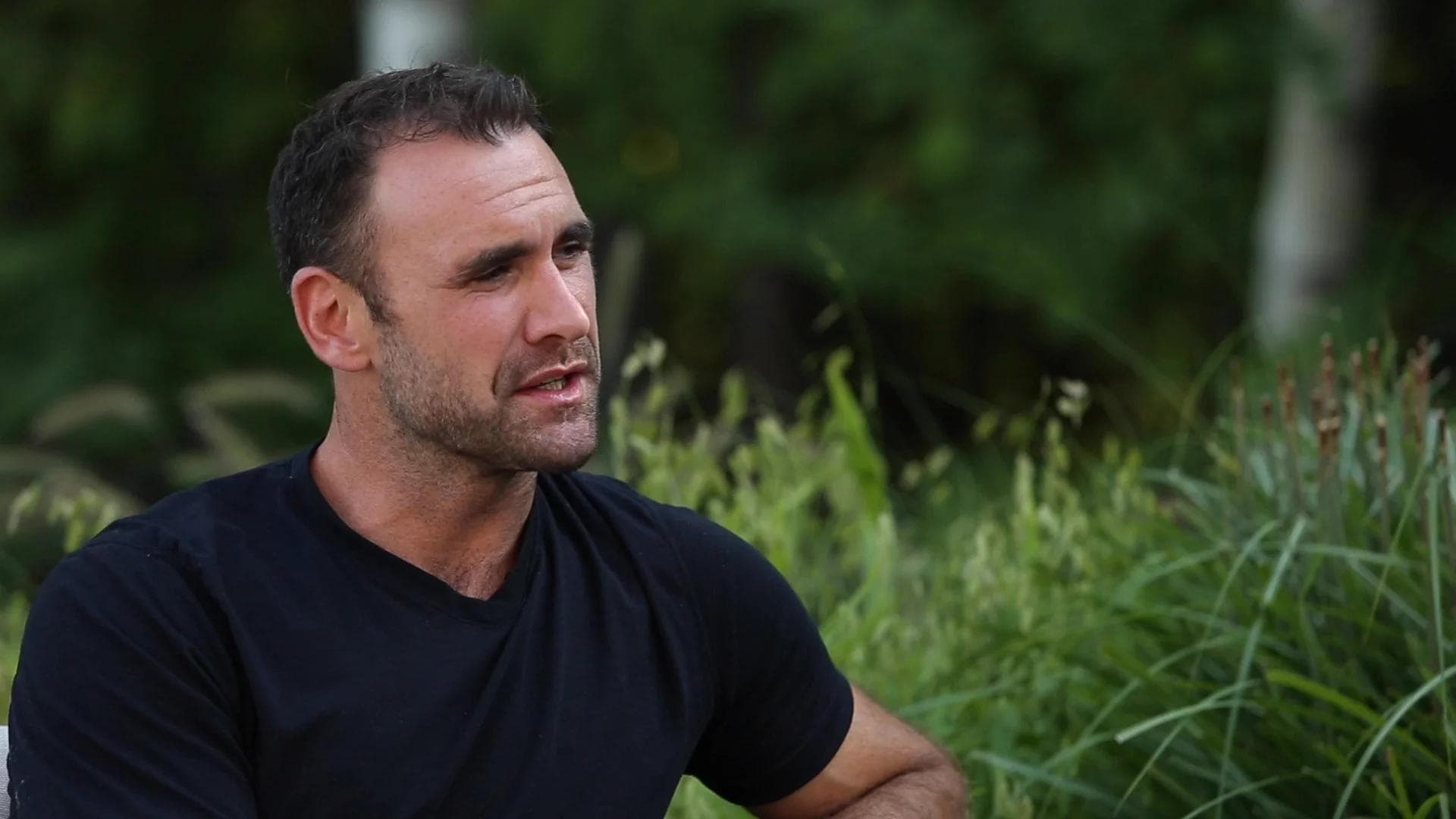Jay Harrison sitting outdoors in a black t-shirt, speaking with greenery in the background.